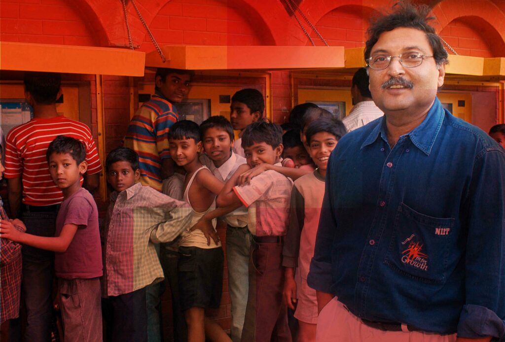 Children line up behind Sugata Mitra to use the hole-in-the-wall PCs installed for independent use. [Image Credit: TED]